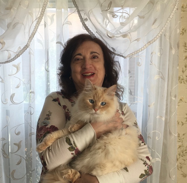 A smiling woman with dark hair holds a fluffy cream-colored cat against a backdrop of sheer white curtains and floral wallpaper.