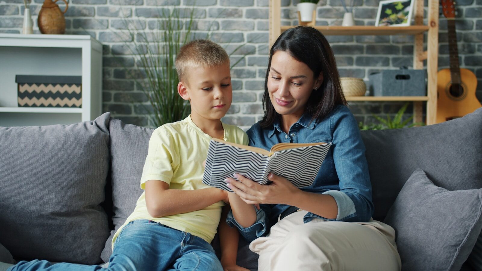 A woman and a boy sit on a couch, reading a book together. A cozy, decorated living room surrounds them, emphasizing quality time and learning.